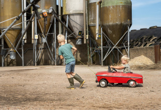 Two children playing at a farm, one pulling the other in a red toy car in front of large farm silos.