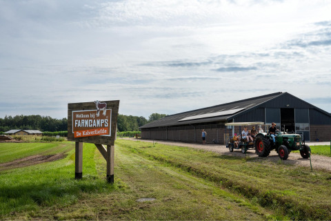 Cartel de bienvenida a FarmCamps De Kalverliefde con tractor y visitantes frente a un granero en Limburg.