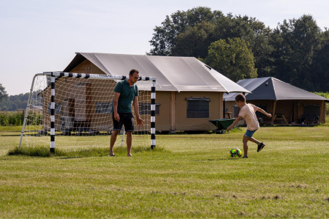 Een man en een kind spelen voetbal op het gras bij tenten op FarmCamps De Kalverliefde in Limburg, Nederland.