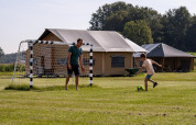 A man and a child play soccer on grass in front of tents at FarmCamps De Kalverliefde, Limburg, Netherlands.