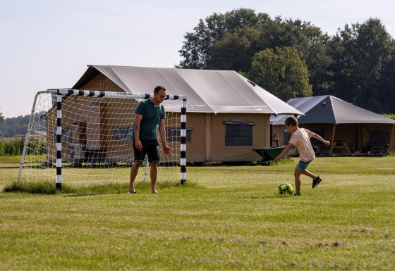 A man and a child play soccer on grass in front of tents at FarmCamps De Kalverliefde, Limburg, Netherlands.