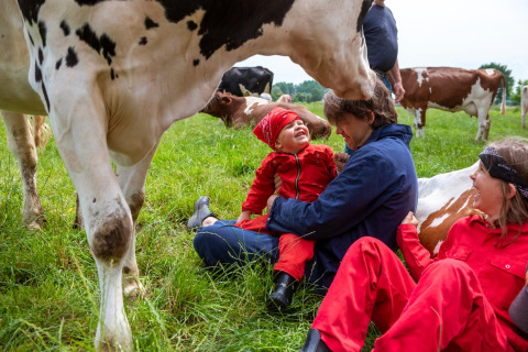 Family in red coveralls sits on the grass laughing with cows at FarmCamps Den Branderhorst, Gelderland, Netherlands.