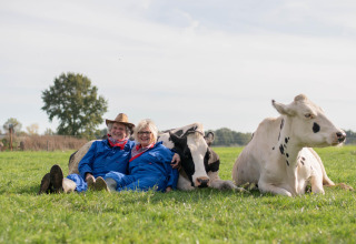 Un couple âgé en salopette bleue assis dans l’herbe à côté de deux vaches à FarmCamps Den Branderhorst.