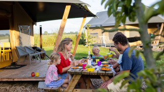 Gezin eet samen aan een picknicktafel bij een safaritent op FarmCamps De Kamperhoek in Zeeland, Nederland.