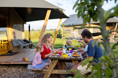 Famille partageant un repas en plein air devant une tente safari à FarmCamps De Kamperhoek, en Zélande.