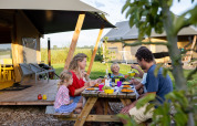 Famiglia che cena all'aperto su un tavolo da picnic davanti a una tenda safari a FarmCamps De Kamperhoek, Zeeland.