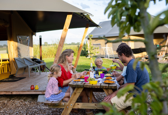 Famille partageant un repas en plein air devant une tente safari à FarmCamps De Kamperhoek, en Zélande.