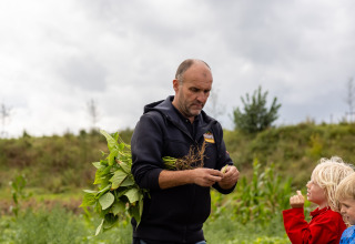 Mand viser børn planter på FarmCamps De Kamperhoek, en feriepark i Zeeland, Holland, under overskyet himmel.
