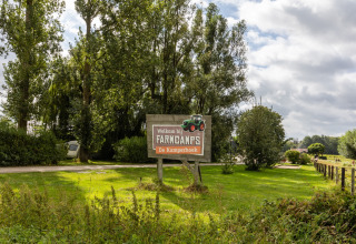 Velkomstskilt ved FarmCamps De Kamperhoek feriepark i Zeeland, Holland, omgivet af grønt og træer.