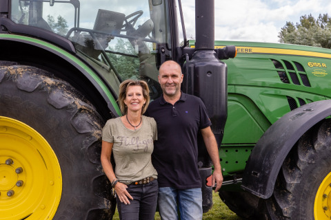 Twee personen poseren voor een grote groene John Deere-tractor bij FarmCamps De Kamperhoek in Zeeland.