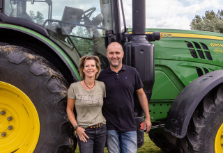 Twee personen poseren voor een grote groene John Deere-tractor bij FarmCamps De Kamperhoek in Zeeland.