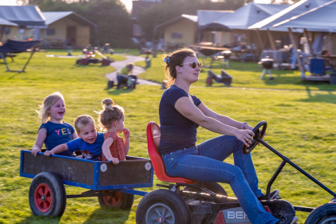 Vrouw rijdt op een tractor met drie blije kinderen in een kar bij FarmCamps De Oostermaat in Overijssel.