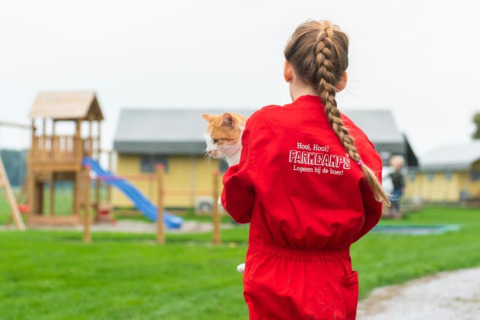Een kind in een rood FarmCamps-pak houdt een kat vast op FarmCamps De Oostermaat in Overijssel, Nederland.
