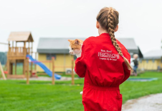 Een kind in een rood FarmCamps-pak houdt een kat vast op FarmCamps De Oostermaat in Overijssel, Nederland.