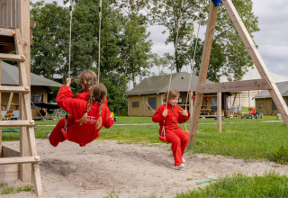 Kinder in roten Overalls schaukeln auf einem Spielplatz bei FarmCamps De Oostermaat, Niederlande.