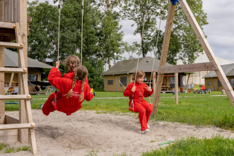 Kinderen in rode pakken schommelen bij FarmCamps De Oostermaat, een vakantiepark in Overijssel.