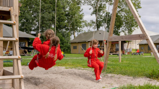 Kinderen in rode pakken schommelen bij FarmCamps De Oostermaat, een vakantiepark in Overijssel.