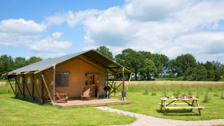 Tienda safari llamada Lodgetent en un campo verde con mesa de picnic bajo cielo azul y nubes.