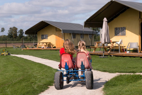 Dos niños en un kart a pedales pasan frente a tiendas en FarmCamps Alpaca Vorstenbosch en Brabante Septentrional.