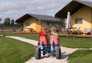 Two kids riding a pedal go-kart past tents at FarmCamps Alpaca Vorstenbosch holiday park in North-Brabant.