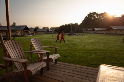 Niños jugando en un campo de césped al atardecer, rodeados de cabañas en FarmCamps Hoeve Sonneclaer, Drenthe.