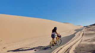 Mujer pedalea en una bicicleta amarilla junto a dunas de arena en TAIGA Tarifa, Andalucía, España.