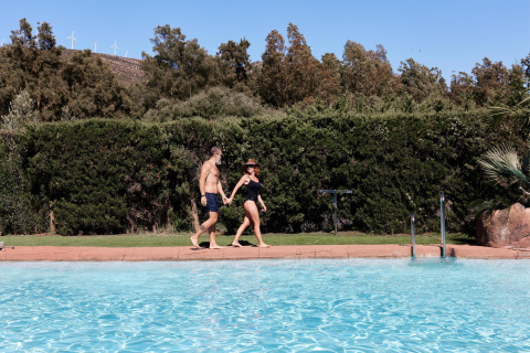 Dos personas en traje de baño caminan de la mano junto a la piscina en TAIGA Tarifa, parque vacacional en Andalucía, España.