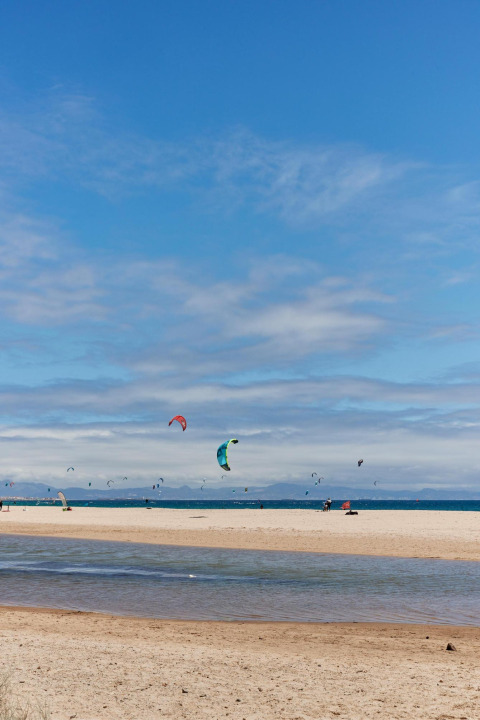 Strand und Kitesurfer bei TAIGA Tarifa, Andalusien, Spanien, unter blauem Himmel mit Wolken.