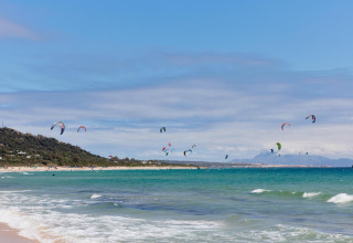 A beach scene with many kitesurfers at TAIGA Tarifa holiday park in Andalusia, Spain, by the turquoise sea.