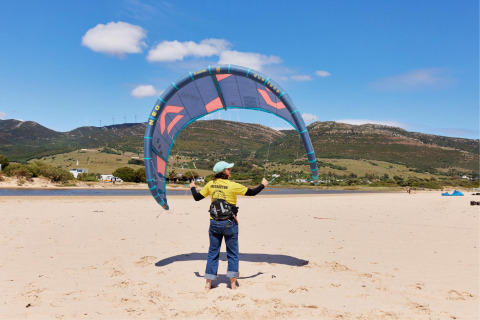 Kitesurf-Instruktor an einem sonnigen Strand bei TAIGA Tarifa Ferienpark, Andalusien, Spanien, mit Bergen im Hintergrund.