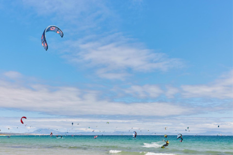 Kitesurfers ride the waves at TAIGA Tarifa holiday park in Andalusia, Spain, with colorful kites in the sky.