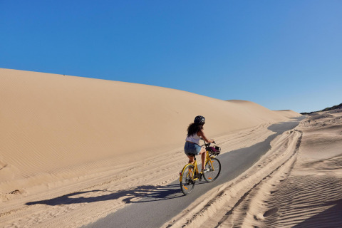 Een vrouw fietst over een pad tussen zandduinen bij TAIGA Tarifa, Andalusië, Spanje, onder een blauwe lucht.