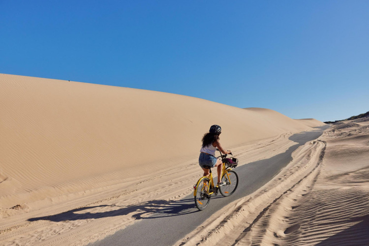 Une femme fait du vélo sur un chemin entouré de dunes et d’un ciel bleu à TAIGA Tarifa, Andalousie, Espagne.