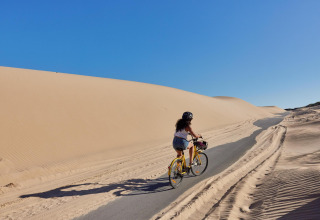 A woman rides a bicycle on a path through sand dunes under a clear sky at TAIGA Tarifa, Andalusia, Spain.