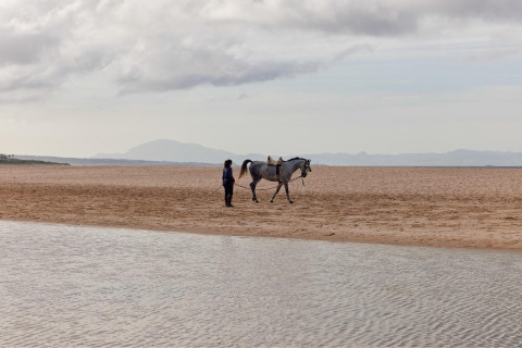 Eine Person führt ein Pferd am Strand des TAIGA Tarifa Ferienparks in Andalusien, Spanien, bei bewölktem Himmel.
