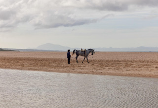 Eine Person führt ein Pferd am Strand des TAIGA Tarifa Ferienparks in Andalusien, Spanien, bei bewölktem Himmel.
