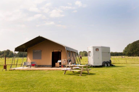 Ranchtent safaritent met wagen, picknicktafel en stoelen op een grasveld onder een blauwe lucht.