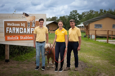 Tres personas y un pequeño poni posan frente al letrero de FarmCamps Holzik Stables en Overijssel, Países Bajos.