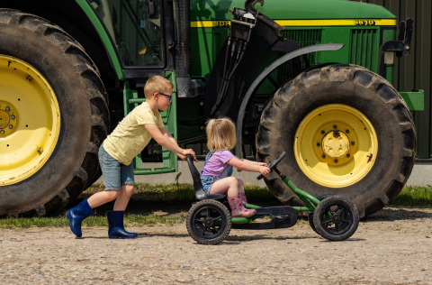 Two kids play with a pedal cart in front of a large green tractor at FarmCamps Holzik Stables, Netherlands.