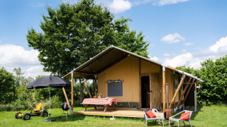 Tente safari avec véranda, table à nappe à carreaux, chaises et jouets dans un cadre verdoyant en plein air.