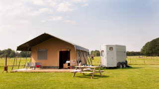 Tienda tipo safari con terraza, mesa de picnic y remolque en un campo verde, perfecta para acampar tranquilo.