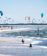 Playa llena de kitesurfistas y cometas, junto a un parque vacacional que ofrece alojamiento glamping.