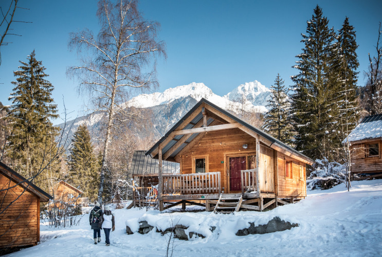 Cabaña de madera en la nieve en Chalet Montana, Les Chalets Huttopia de Bozel, Francia, con montañas al fondo.