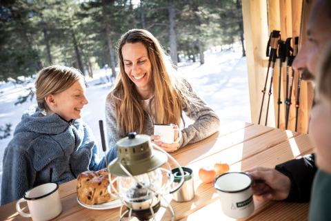 Familie genießt Frühstück und heiße Getränke in einer Holzhütte im Schnee in Südfrankreich.