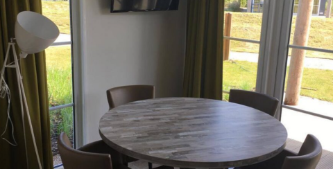 Ronde houten tafel met vier stoelen, vloerlamp en grote ramen in Comfort Lodge op Kompas Camping Nieuwpoort, België.