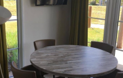 Ronde houten tafel met vier stoelen, vloerlamp en grote ramen in Comfort Lodge op Kompas Camping Nieuwpoort, België.
