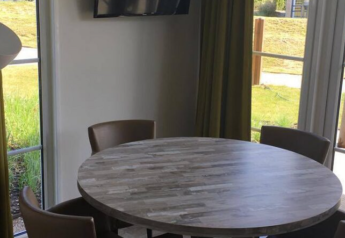 Tavolo rotondo in legno con quattro sedie, lampada da terra e grandi finestre presso Comfort Lodge, Kompas Camping Nieuwpoort, Belgio.