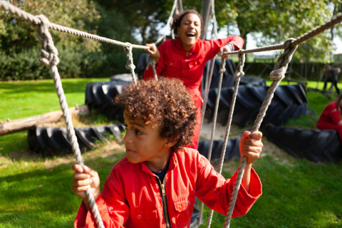 Children in red coveralls playing on a rope bridge outdoors at FarmCamps Hoeve Sonneclaer in Drenthe, Netherlands.