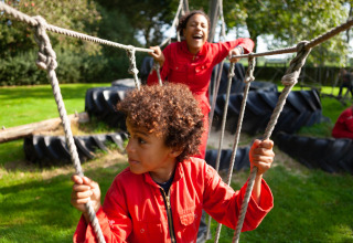 Children in red coveralls playing on a rope bridge outdoors at FarmCamps Hoeve Sonneclaer in Drenthe, Netherlands.