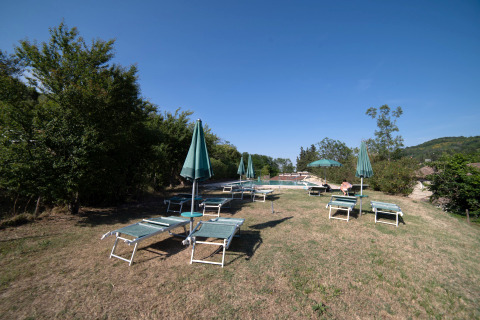 Solstole og parasoller på en tør græsplæne ved Camping Podere Sei Poorte, Marche, Italien, under en klar himmel.